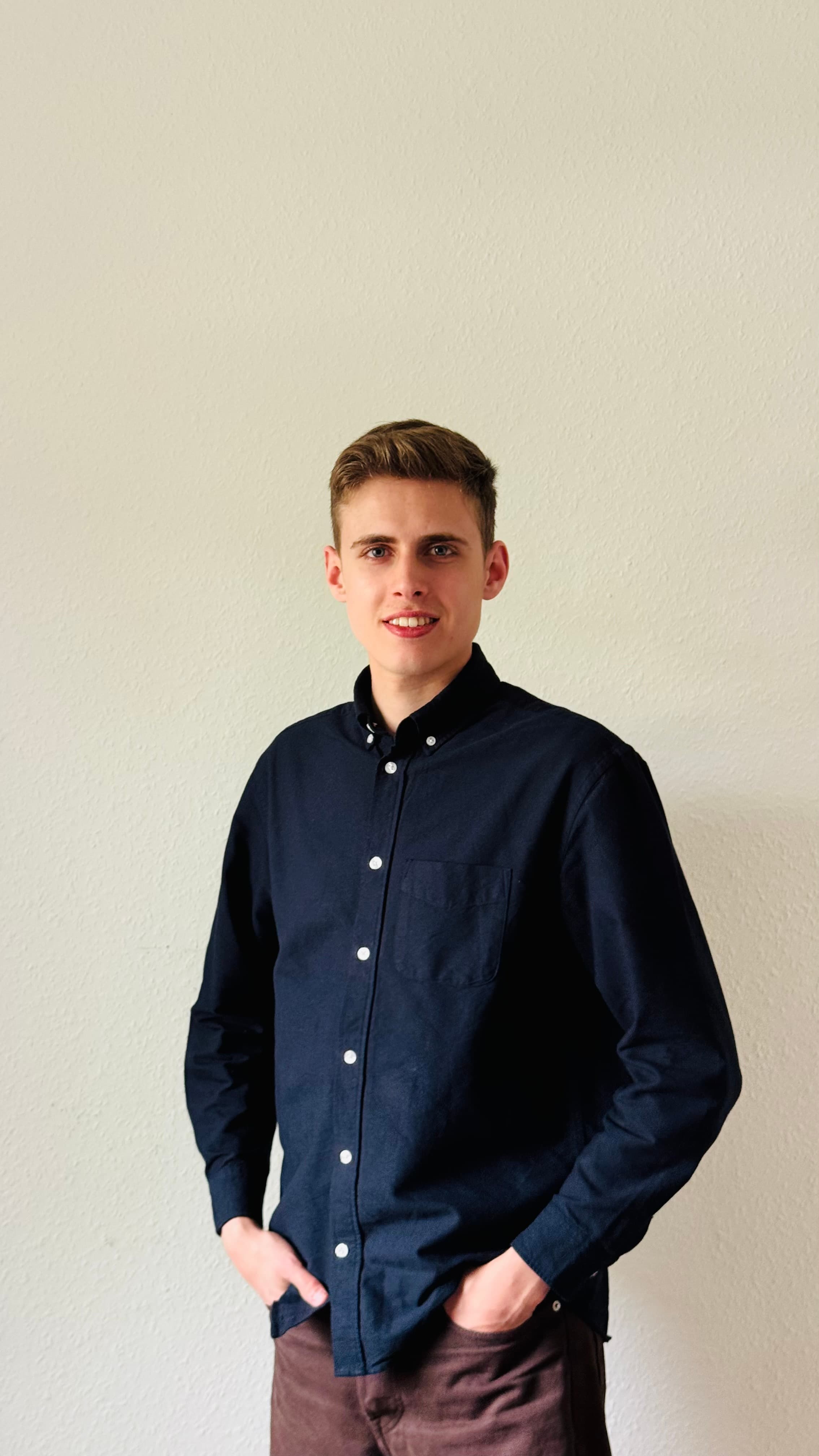 Lächelnder junger Mann im dunkelblauen Hemd steht mit Händen in den Taschen vor weißer Wand.