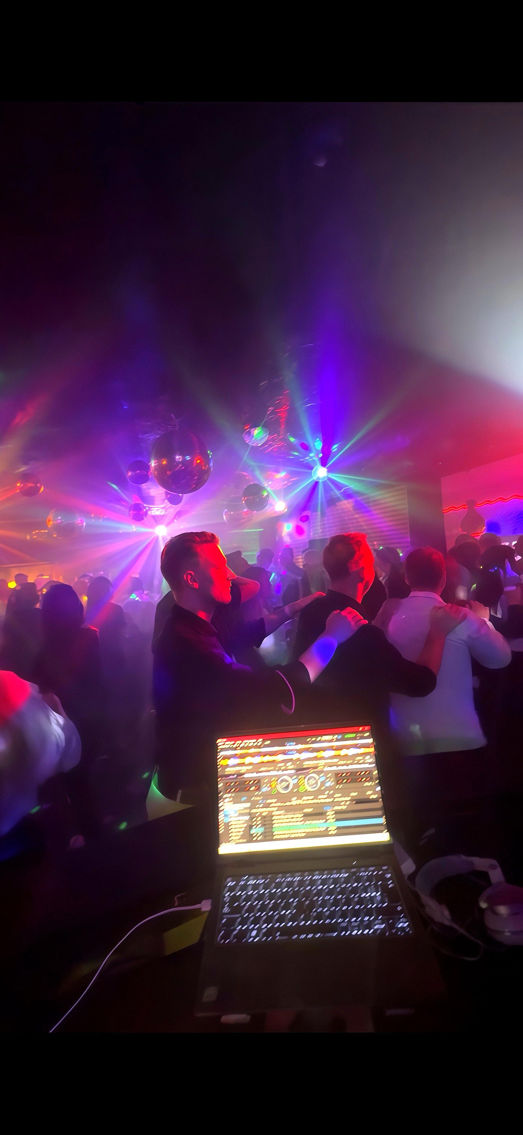 DJ-Laptop im Vordergrund, tanzende Menschen unter bunten Laserlichtern und Discokugeln in einem dunklen Nachtclub.