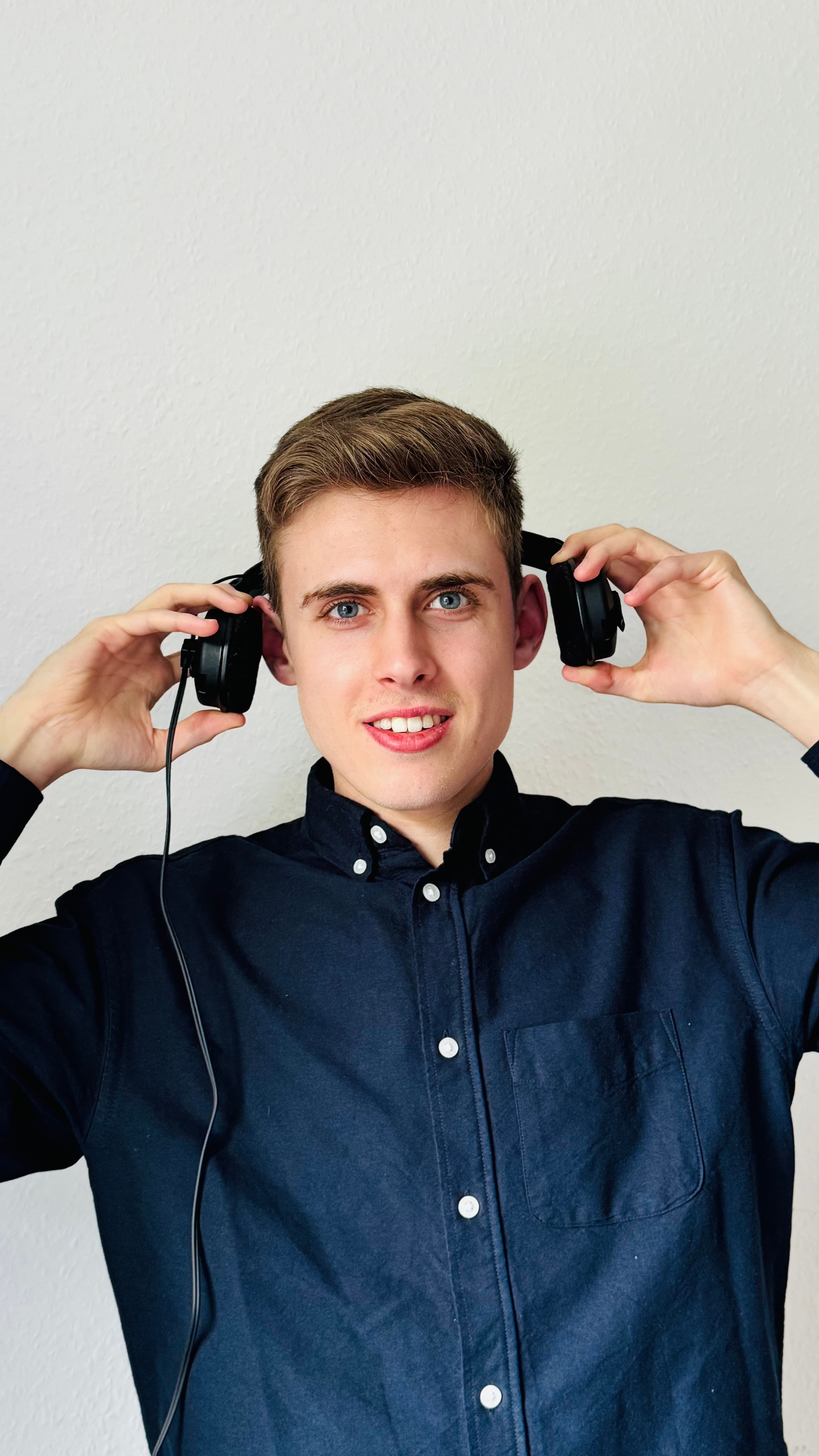 Junger Mann mit blauen Augen im blauen Hemd hält sich schwarze Kopfhörer an die Ohren.